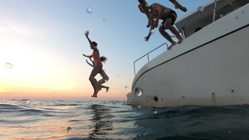 Group of young friends jumping to the beach, having a party in yacht. Attractive man and woman people hanging out, celebrating holiday vacation trip while catamaran boat sailing during summer sunset.