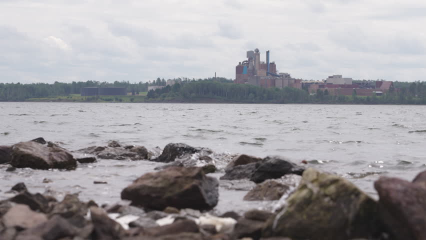 From a rocky shoreline, a factory is seen across the lake, on a windy overcast day
