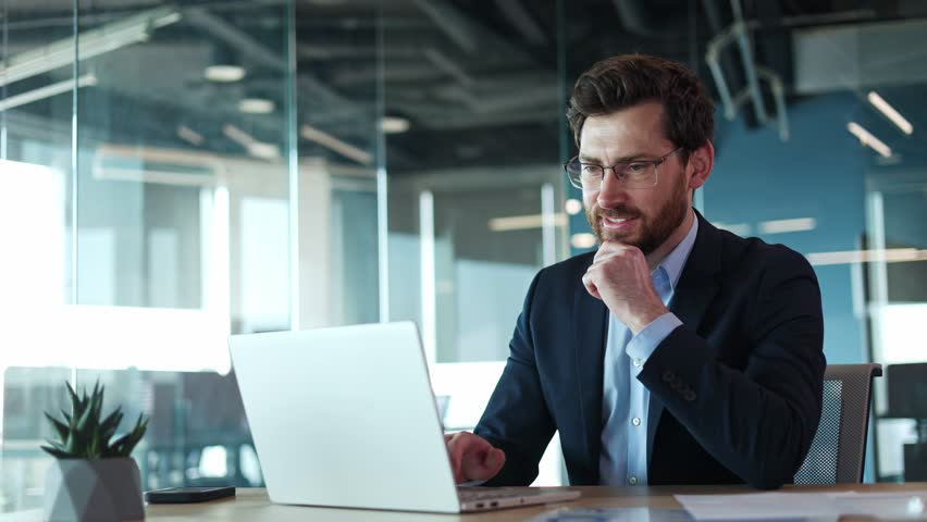 Smiling caucasian male wearing glasses with protective coating and typing on portable computer on background of office environment. Cheerful young businessman doing favorite work with pleasure.