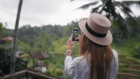 Blonde woman records video on sea beach. Tourist female on resort admiring nature shooting seascape on smartphone at sunset, rear view. Tourism, travel, create content, admiring vacations concept.