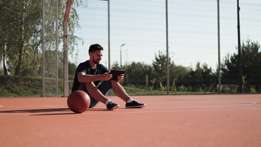 Caucasian man during break sits with ball on basketball court and taps on tablet. Watch online broadcast of important game on sunny day. Using mobile Internet outdoors for entertainment.