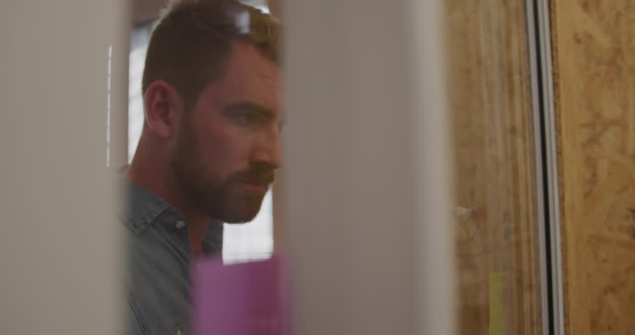Side view of young Caucasian creative professional man in a modern office writing on a mood board full of sticky notes