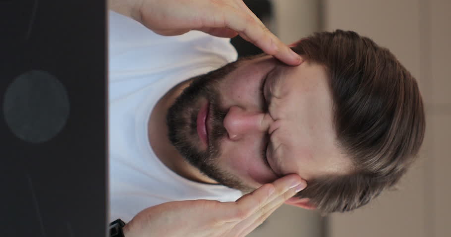 Vertical video: Man holding hands, massaging forehead and head with severe pain. Freelancer man work from home kitchen suffer with headache. Hard studying, busy student, sad.