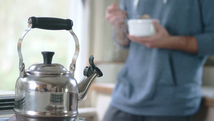 Tea kettle steaming on stove top while young man eats cereal in background