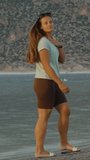 Vertical video. A young woman bathed in the sunset's glow strolls along the shore of Mountain Lake and points into the distance. The camera quickly turns in that direction.