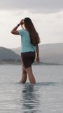 Vertical video. Clouds and gray sky over the mountain lake, Sun rays struggling to break through the clouds. A Young Woman walks on the water.