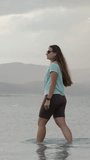 Vertical video. Approaching Storm Clouds in the distance near the mountains. They are moving towards the lake where a Young Woman is walking in the water and pointing at them.