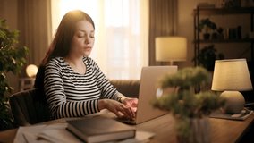 Woman working on laptop at home while talking on phone, backlit warm light