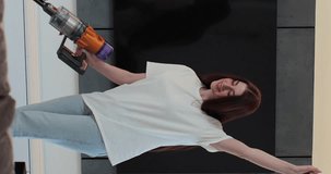 Vertical video: Woman dancing with a vacuum cleaner at home. Young woman having fun while cleaning house. House cleaning, housework concept. Smart cleaning technology.