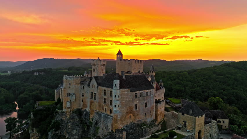 Cinematic panoramic view of feudal square-shaped castle on a riverside clifftop at golden sunset in France. Aerial view of Château de Beynac fortress at golden hour in France.