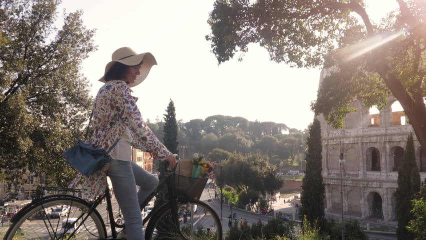 Beautiful young woman in colorful fashion riding bike in front of colosseum in Rome at sunset with trees attractive girl with straw hat steadycam dolly