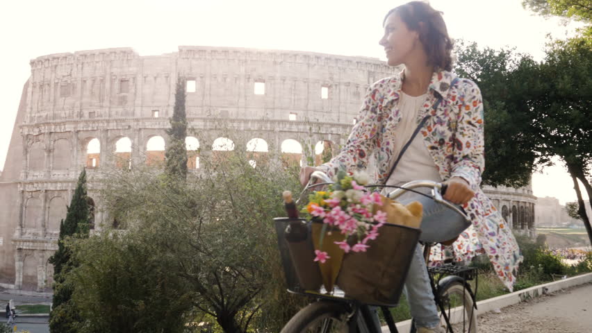 Beautiful young woman in colorful fashion riding bike in front of colosseum in Rome at sunset with trees attractive girl with straw hat front view steadycam dolly