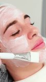 Vertical video. A cosmetologist applies anesthetic cream to a young woman's face before a facial cosmetic procedure. Anesthetic cream. A preparation for healing, tightening, and rejuvenating skin