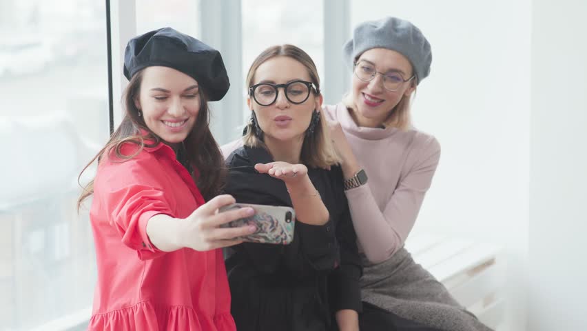 Three young stylish girlfriends spend time together, take photos on a smartphone and look at them. Three young women are smiling, making selfies. Girls in beret, glasses. Stylish girlfriends. | Shutterstock HD Video #34784119