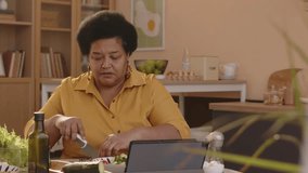 Stab waist up shot of adult Black woman having video chat on tablet pc while making lunch at home, cutting fresh vegetables for salad at cooking table in cozy kitchen