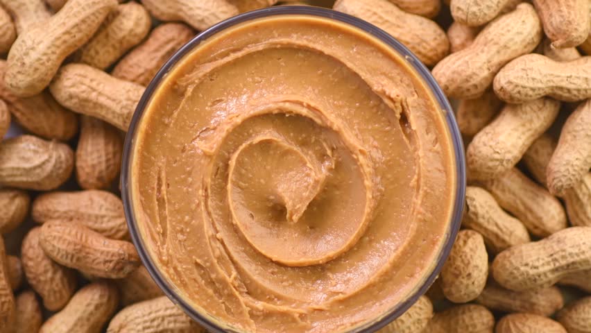 Peanut butter in a glass bowl over raw peanuts background. Creamy smooth peanut butter in glass bowl backdrop. Texture. Organic food. American cuisine. top view