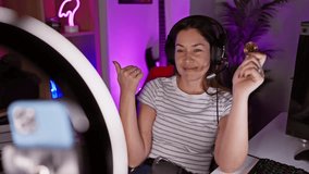 Bubbly young woman with blue eyes records bitcoin video in gaming room, pointing thumb to the side with a big, cheery smile and open mouth