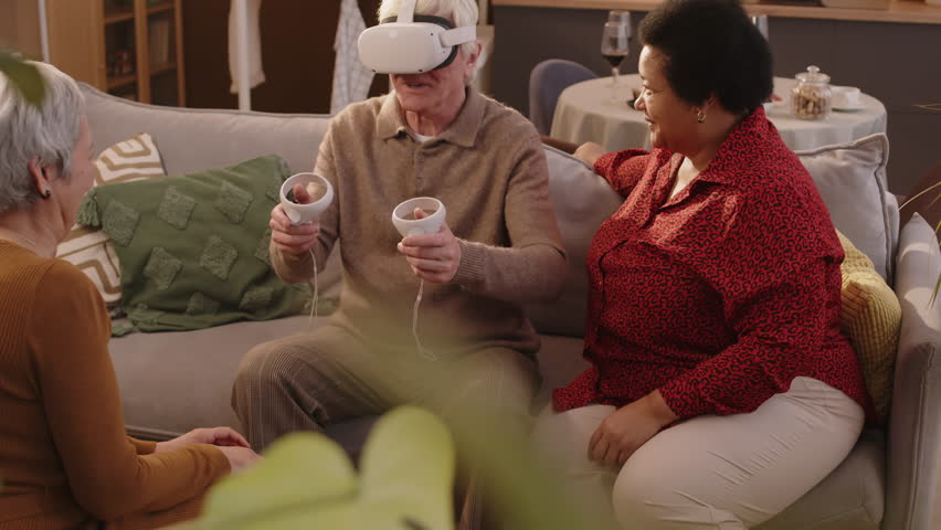 Medium shot of Caucasian aged man in vr headset playing vr game while getting together with two old diverse female friends in bright cozy apartment Royalty-Free Stock Footage #3478523761