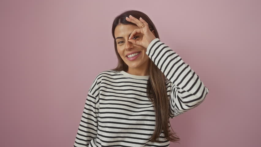 Happy young hispanic woman in stripes t-shirt standing joyfully, confidently flashing a smile and ok sign over her eye, peering through fingers on isolated pink background.