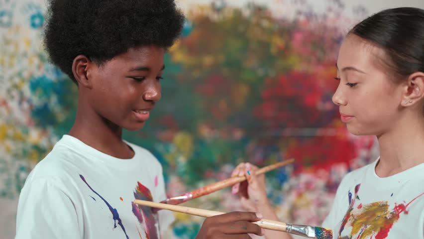 Smiling diverse children using paint brush painted color on each other white shirt shirt at colorful stained wall in art lesson. Represent exchanging experience, learning each other. Edification
