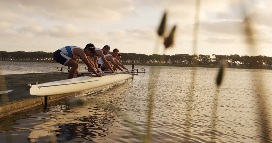 Side view of four Caucasian male rowers, standing in a row on a jetty, holding a boat, taking it out of the water, holding it above their heads bottom up, during a sunset, in slow motion