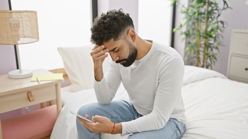 A worried young hispanic man with a beard sits on a bed, holding a smartphone in a modern bedroom.