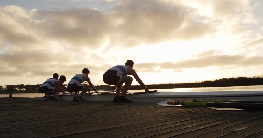 Rear view of four Caucasian male rowers, standing in a row on a jetty, holding a boat, taking it out of the water, holding it above their heads bottom up, during a sunset, in slow motion
