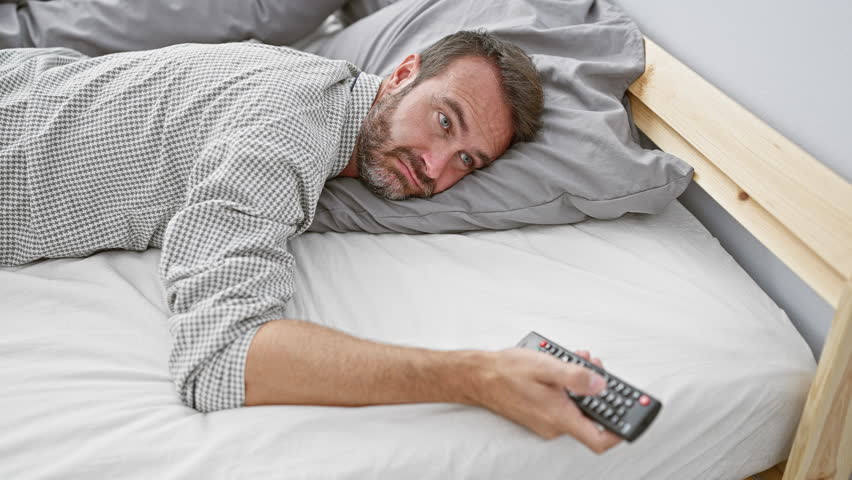 Middle-aged bearded man lounging in bed with a remote control in a cozy bedroom setting