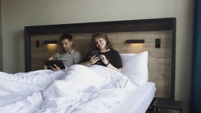 Man and woman are lying in bed under blanket and looking at their phone and tablet. Married couple smilingly watches entertaining videos and reading ebook on weekend morning in their bedroom. - Powered by Shutterstock - Get 15% off with code: PIKWIZARD15