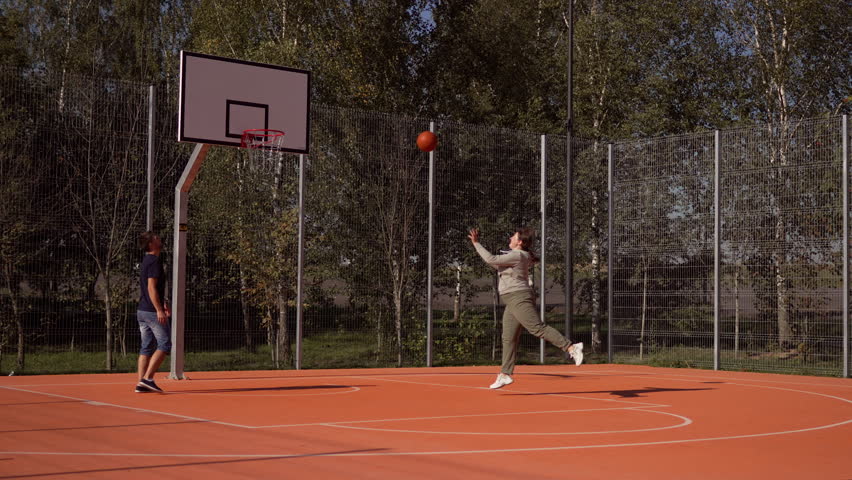 On basketball court man throws ball into basket after woman misses. Playing basketball together on street court and helping you achieve your goal. Combination training with pass to partner.