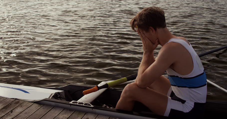 Side view of a Caucasian male rower, after a rowing practise, sitting in a boat by a jetty, hiding his face in his hands, on a sunny day, in slow motion