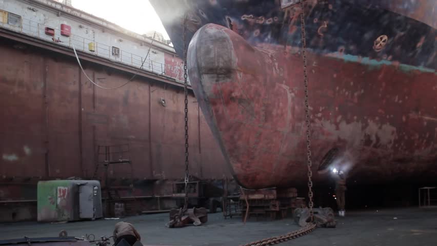 Workers repair ship hull in dry dock. Welding, grinding performed on massive vessels bow. Industrial maritime maintenance, shipbuilding process captured. Rust, repair tools, visible in shipyard.