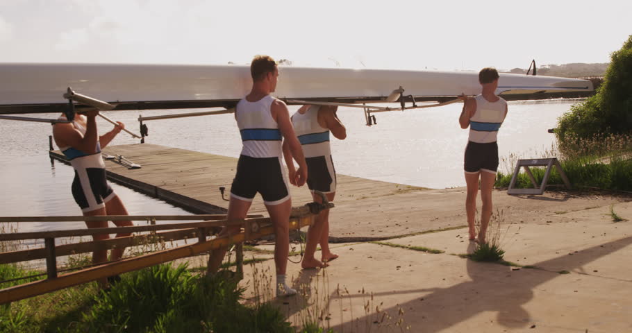 Side view of four Caucasian male rowers, standing in a row on a jetty, holding a boat on their shoulders, walking with it, on a sunny day, in slow motion
