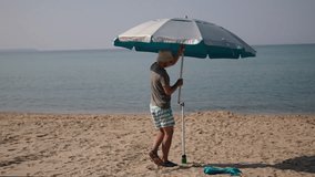 Caucasian tanned man unfolds and sets up large beach umbrella on sea beach on sunny day. Arrangement of place to relax and protection from sun while relaxing on ocean coast during tourist season. - Powered by Shutterstock - Get 15% off with code: PIKWIZARD15