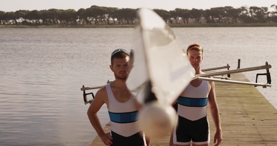 Front view of four Caucasian male rowers, standing in a row on a jetty, holding a boat on their shoulders, walking with it, on a sunny day, in slow motion