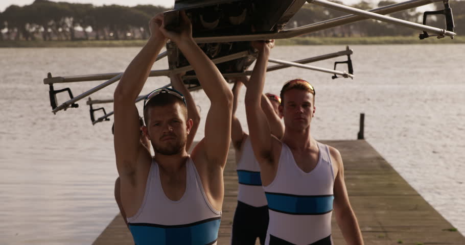 Side view close up of four Caucasian male rowers, standing in a row on a jetty, holding a boat above their heads bottom up, putting it on their shoulders, during a sunset, in slow motion