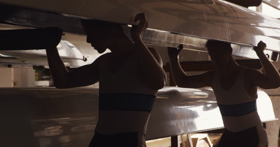 Side view of four Caucasian male rowers, standing in a row in a boathouse, holding a boat above their heads, putting it on a shelves, on a sunny day, in slow motion