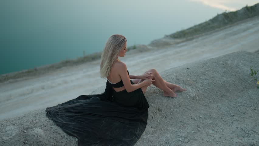 Woman in black dress sits barefoot on shore of lake in evening. Flirting and seduction concept.