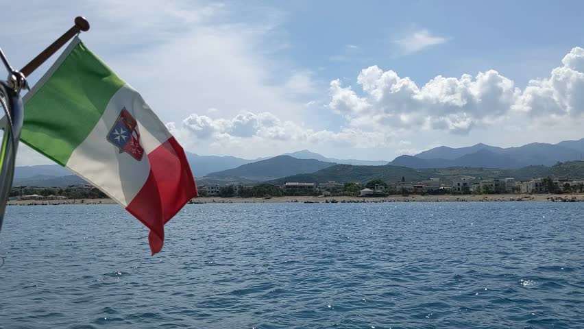An Italian tri-color with a shield representing the four Maritime Republics. Above the shield is a naval crown. Beach view in Sicily, Italy. Yachting in summer vacation. Coast, mountains, cloudy sky - Powered by Shutterstock - Get 15% off with code: PIKWIZARD15