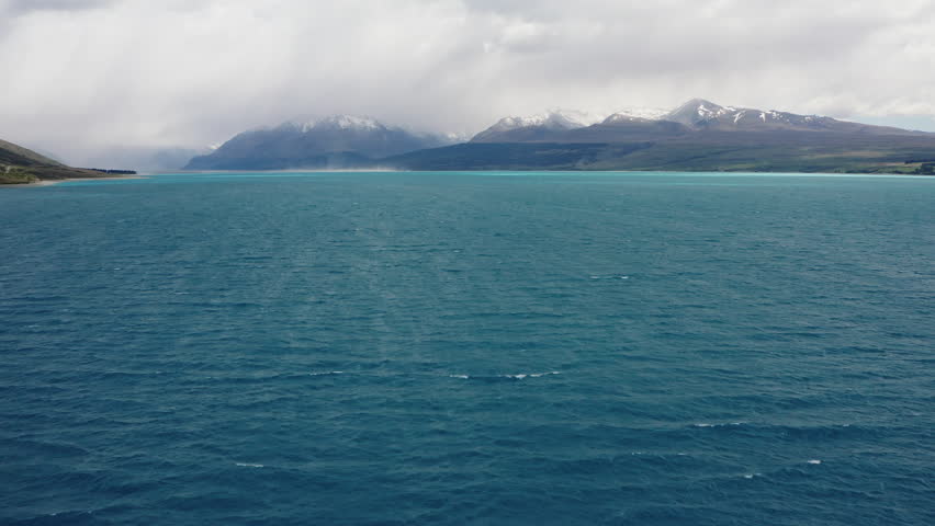 Fly over bright blue water on Lake Pukaki while low clouds shroud the surrounding snow capped picturesque mountains.