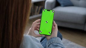 Girl using smartphone with green screen in living room watching movie, video content. Tracking markers. Indoors. Browsing social network, news. Internet surfing communication online. Scroll up closeup