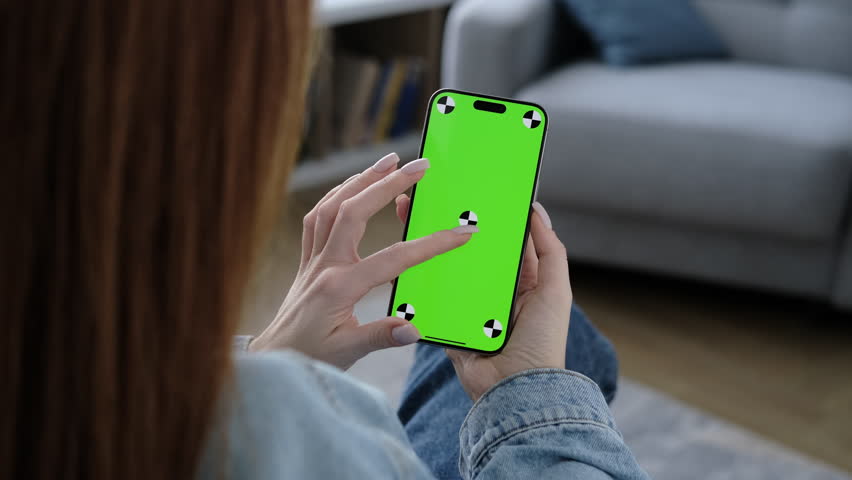 Girl using smartphone with green screen in living room watching movie, video content. Tracking markers. Indoors. Browsing social network, news. Internet surfing communication online. Scroll up closeup