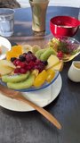 A refreshing beachside breakfast in stunning vertical video. Delight in a colourful fruit bowl with kiwi, red fruits, oranges, and apples. Bask in the sunlight toast with jam, honey and butter.