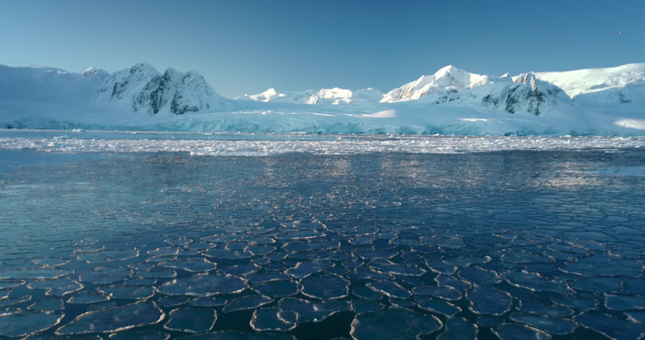 Aerial view ice polar ocean and snow covered mountains in background. Blue clear sky in sunny winter day. Fly over the untouched wilderness of Antarctica. Polar frozen ocean landscape. Travel, explore