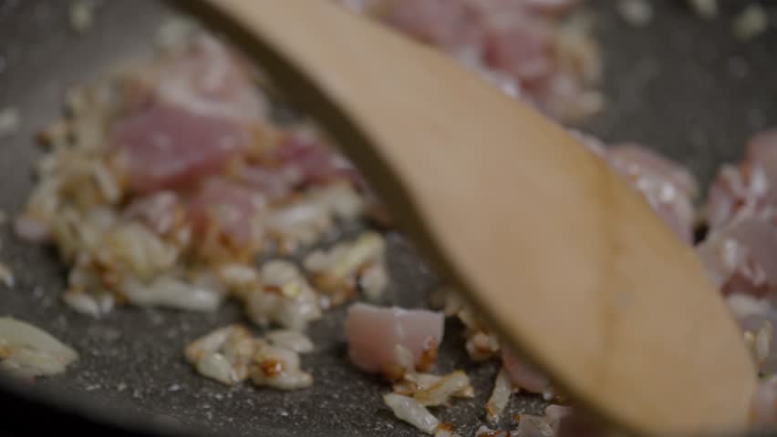 The cook carefully mixes fried onions with a golden brown color and chicken fillets in a black ceramic frying pan using a wooden spatula. Extreme close-up. Slow motion. 4k