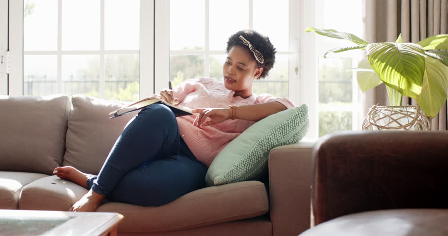African American woman lounges on a sofa with a book at home. Sunlight filters through large windows, creating a cozy, relaxed home atmosphere, slow motion. Royalty-Free Stock Footage #3478889939