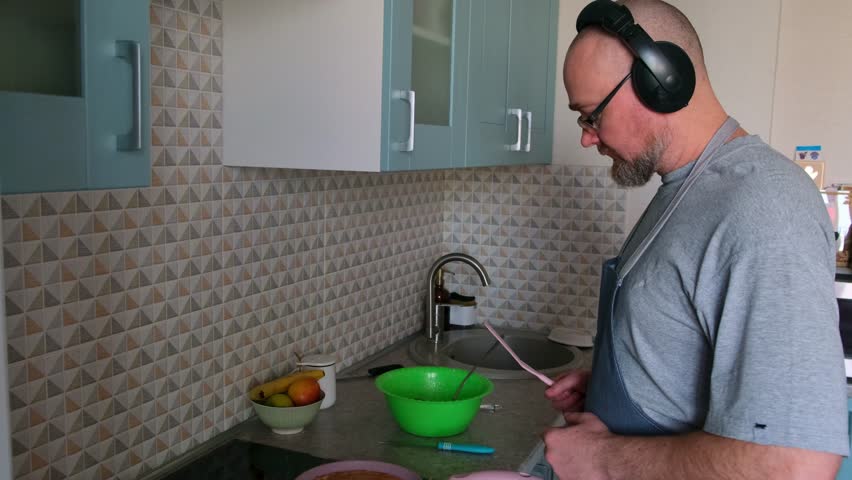 Adult brutal man cooking breakfast in the home kitchen and listening music in headphones. Bald bearded guy wearing apron having fun while baking pancakes on the stove. Royalty-Free Stock Footage #3478915845