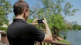 Man capturing landscape photo with smartphone from bridge overlooking tranquil lake surrounded by lush greenery. Technology and leisure.