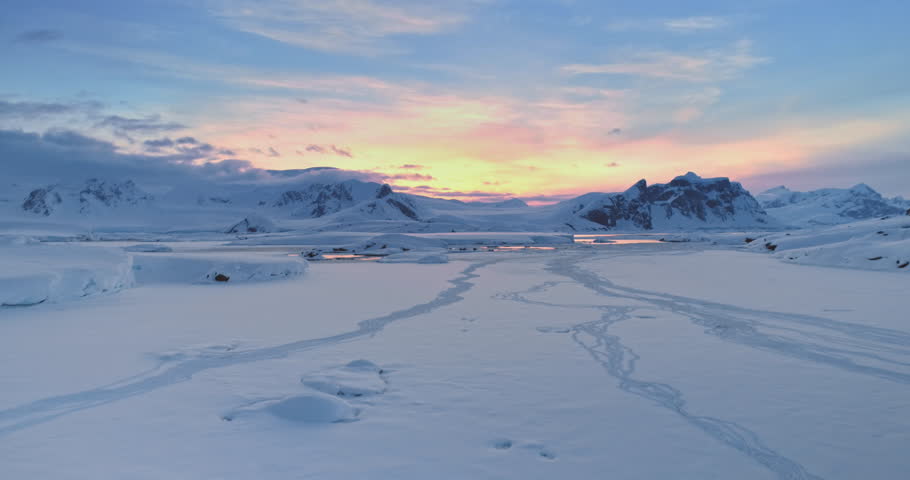 Amazing sunset over snowy Antarctic landscape. Snow covered frozen polar ocean and mountain range under colorful sunrise sky. Arctic winter nature. Antarctica travel and exploration. Aerial drone shot