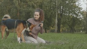 Young joyful Caucasian woman taking selfie photo or recording video with her lovely mixed breed dog, resting together on green grass in park at daytime